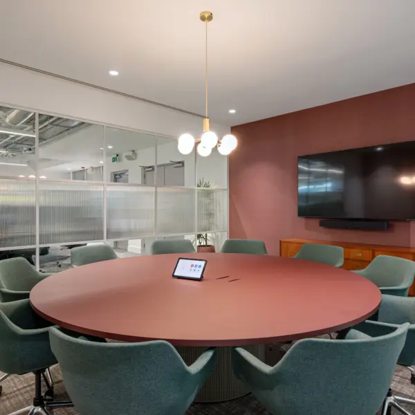 Contemporary meeting room with a round, muted red table at the centre, surrounded by eight upholstered swivel chairs in a soft green tone on castors. A small touchscreen meeting control panel sits in the middle of the table. Above, a pendant light with multiple spherical bulbs hangs from the ceiling, providing warm, even illumination.  The right-hand wall is finished in a textured, deep red surface and features a large wall-mounted flat screen with a soundbar below, positioned above a low wooden sideboard. To the left, a glass partition wall with frosted lower panels and clear upper sections separates the room from the open office beyond, where desks, chairs and exposed ceiling services are visible.  The space is finished with neutral carpeting, white ceilings with recessed lighting and clean-lined detailing throughout. A small potted plant sits near the glass wall, adding greenery to the otherwise structured, modern setting.