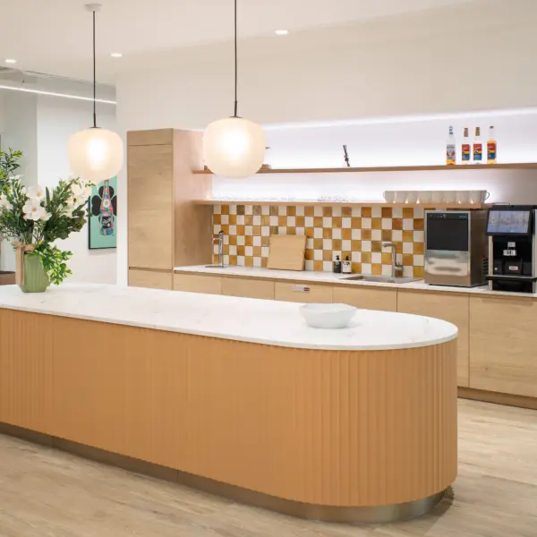Modern office kitchenette and breakout space with a clean, minimalist design. In the foreground, a large curved island counter with a fluted wood front and a white stone worktop sits on light wood flooring, with a small bowl and a tall vase of flowers placed on top. Two round pendant lights hang above the island, casting soft, even light.  Behind the island, a wall of light wood cabinetry integrates appliances including a built-in oven and a coffee machine. Open shelves display glassware, mugs and bottles, while a warm-toned, checkered tile backsplash adds visual interest. A sink and preparation area run along the counter beneath the shelving.  To the left, the space opens towards the wider office, with a glimpse of glass partitions, additional greenery and wall art. The overall palette combines light timber, white surfaces and warm accents, creating an inviting, contemporary feel.