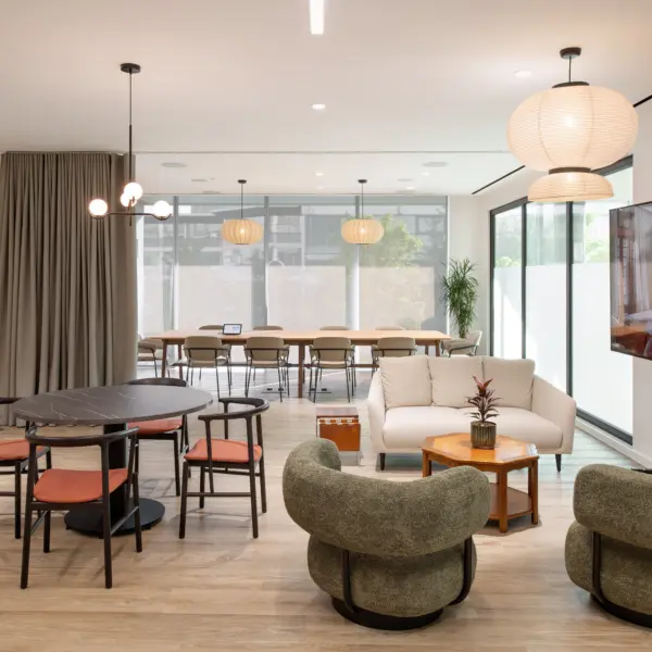 Modern breakout and collaboration area within an office, combining lounge seating and shared workspaces. In the foreground, two rounded upholstered armchairs in a muted green tone face a small wooden coffee table with a plant on top, alongside a light-coloured sofa and an adjacent side table. To the left, a circular dark table is surrounded by four chairs with wooden frames and muted red seat pads, creating a more casual meeting spot.  To the right, a large freestanding digital display screen sits on a stand, positioned near the seating area. Above, a mix of pendant lights hangs from the ceiling, including sculptural multi-globe fixtures and larger paper-style lantern shades, adding soft, warm lighting.  In the background, a long communal table with multiple chairs sits in front of full-height windows with frosted lower sections, allowing natural light to fill the space while maintaining privacy. Floor-to-ceiling curtains line the left side, and a tall potted plant adds greenery near the edge of the frame.  The space features light wood flooring, white walls and a clean, contemporary finish, blending informal seating with functional work areas for meetings or relaxation.