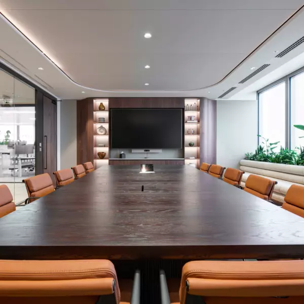 A modern conference room featuring a large wooden table surrounded by brown leather chairs. One side of the room is enclosed with glass walls, while the far end features a wall-mounted flat-screen television and built-in shelving displaying decorative items. Large windows along one side let in natural light and are adorned with potted plants on the sills, adding a touch of greenery to the professional setting.