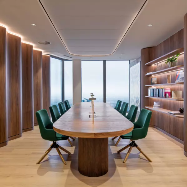 A modern conference room featuring a long wooden table surrounded by green upholstered chairs. The walls are clad in wooden panelling, with built-in shelving units displaying books and decorative objects. Large windows at the far end of the room allow natural light to flood the space. The ceiling is fitted with recessed lighting, contributing to the room’s bright and welcoming ambience.