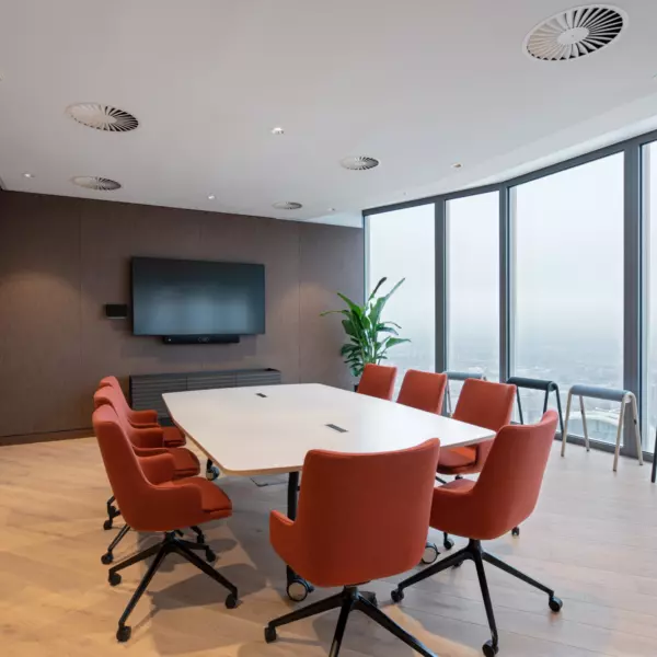 A modern conference room featuring a large white table surrounded by eight orange swivel chairs. Floor-to-ceiling windows provide expansive views of the city skyline, including a prominent skyscraper in the distance. The room includes a wall-mounted flat-screen television and a whiteboard on an adjacent wall. Wooden flooring and ceiling lights contribute to the clean, professional atmosphere. Three high stools are positioned near the windows, and a green plant in the corner adds a touch of nature.