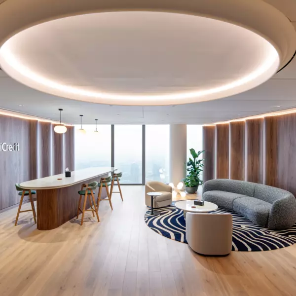 A stylish and modern office lounge area featuring a curved grey sofa that forms a semi-enclosed booth-like seating arrangement around a round coffee table placed on a patterned rug. The space includes a sleek oval-shaped bar table with high stools, positioned under a large circular ceiling light fixture. Wooden flooring and wall paneling add warmth to the environment, while a tall indoor plant in the corner and large windows offering natural light enhance the inviting atmosphere. The word "UniCredit" is displayed on the wall to the left, contributing to the branded interior design.
