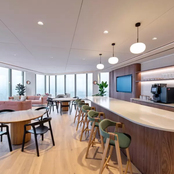 A contemporary office lounge area with large windows offering an outside view and allowing natural light to brighten the space. The room features several round tables with chairs arranged for casual seating, alongside a long counter fitted with bar stools. A coffee machine is positioned on the counter, suggesting a refreshment area. Additional elements include potted plants for a touch of greenery and a wall-mounted television screen for information or entertainment.