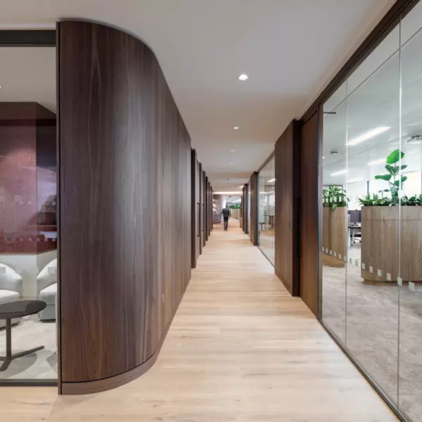 A contemporary office corridor with wooden flooring and wall panelling. On the left, glass-walled meeting rooms are visible, one containing white chairs and a small table. To the right, glass partitions separate the hallway from an open-plan office area furnished with desks and decorated with green plants. The space is brightly lit with ceiling lights, and a person is seen walking at the far end of the corridor.
