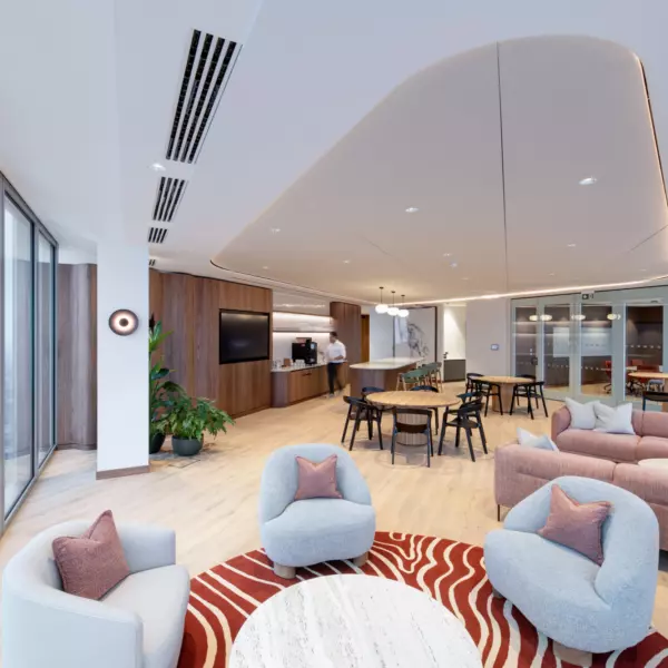 A modern, open-plan living space with large windows along the left side, allowing natural light to fill the room. The foreground features a seating area with three light grey armchairs arranged around a round white marble coffee table, set atop a red and white patterned rug. In the background, a dining area with several tables and chairs leads into a kitchen or bar area fitted with wooden cabinetry. The ceiling includes recessed lighting and a distinctive architectural design, while potted plants near the windows add a touch of greenery.