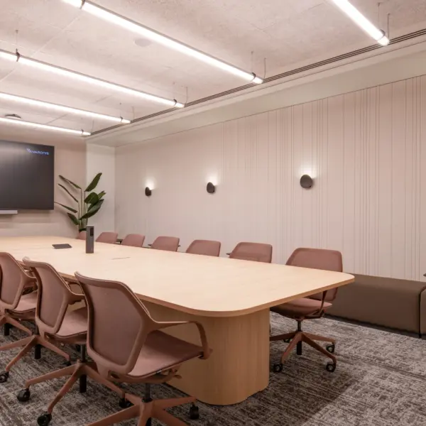 Contemporary boardroom featuring a large light oak conference table surrounded by ergonomic meeting chairs in a muted terracotta tone. A wall-mounted display screen sits at one end of the room, while acoustic wall panelling, integrated lighting and neutral finishes create a clean, professional meeting environment. A built-in bench seat and planting add warmth and comfort to the space.