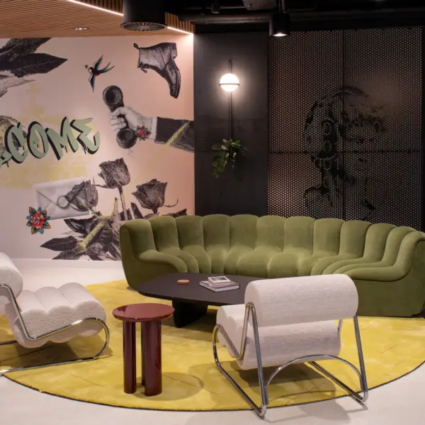 Contemporary office lounge area featuring a sculptural green curved sofa, two white accent chairs and a black coffee table arranged on a circular yellow rug. A large feature mural with mixed-media artwork and the word "WELCOME" forms the backdrop, while dark walls, timber slatted ceiling details, decorative lighting, planting and colourful statement pieces create a bold and inviting reception space.