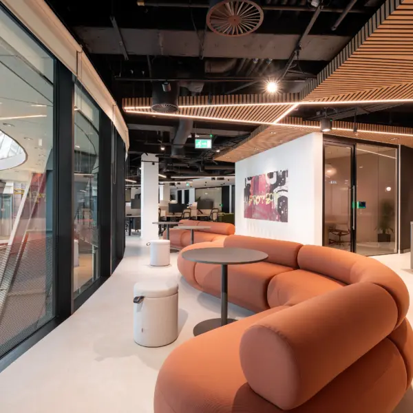 Modern office lounge area featuring a curved terracotta-coloured sofa and round tables positioned alongside floor-to-ceiling glazing. The space leads into an open-plan workplace with meeting rooms, collaborative seating and a reception area displaying Varonis branding. Timber slatted ceiling features, exposed black services and warm lighting create a contemporary, high-specification office environment.
