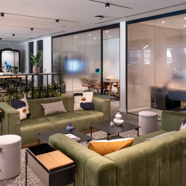 Contemporary office breakout and collaboration area featuring two green sofas arranged around black coffee tables, with adjacent informal seating and meeting spaces. Floor-to-ceiling glass partition walls enclose meeting rooms, while planters, soft furnishings and modern furniture create a comfortable workspace with a neutral colour palette and abundant natural light.