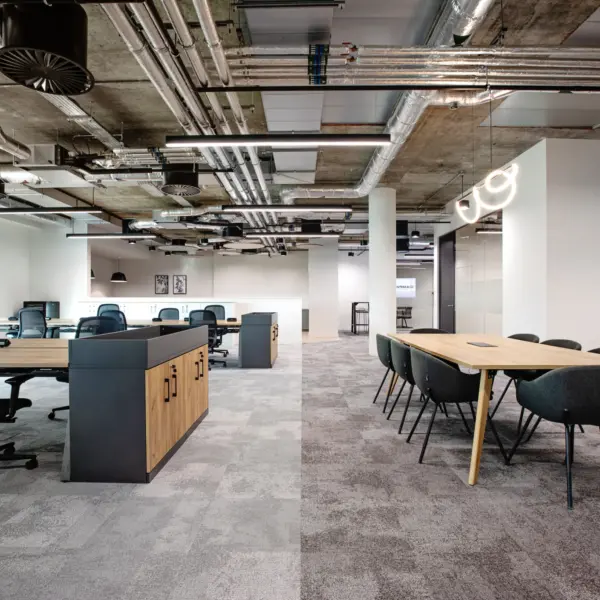 Open plan office with a contemporary industrial finish, featuring shared workstations and a meeting area. In the foreground on the right, a rectangular light wood meeting table is surrounded by dark upholstered chairs with slim black legs. To the left, rows of desks with light wood tops and black frames are paired with black mesh swivel chairs, separated by low storage units with wood fronts and dark sides.  The space extends into the background with more desks and chairs arranged in an orderly layout. A white partition on the right includes a doorway with a glowing “go” sign above it. The ceiling is exposed, showing silver ductwork, pipework and suspended black acoustic discs alongside linear lighting. Large windows on the left bring in natural light, while grey carpet flooring runs throughout. The space is clean, minimalist and unoccupied.