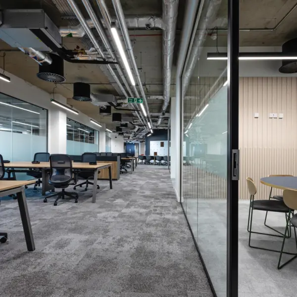 Open plan office interior with a contemporary, industrial finish. Rows of light wood desks paired with black mesh swivel chairs sit on a grey carpet in the foreground and extend into the background. To the right, a glass partition and door separate a small meeting area with a round table and upholstered chairs against a light wood panelled wall. Exposed ceiling services including silver ventilation ducts, pipes and linear strip lighting run overhead. Frosted glass panels line the left side, allowing light to pass between spaces while maintaining privacy. The space is clean, minimal and unoccupied.