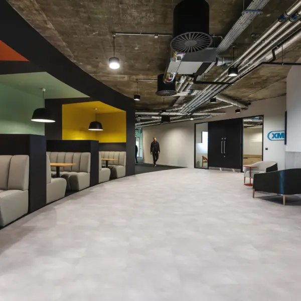 Modern office breakout area with an open industrial ceiling featuring exposed ductwork and lighting. On the left, a row of upholstered booth seating in neutral tones lines the wall, each booth paired with a small round wooden table and set within colourful alcoves in shades of green, yellow and orange. Pendant lights hang above the seating. The centre of the space has a wide, light grey floor leading towards a corridor, where one person is walking in the distance. On the right, there is a lounge area with a cushioned chair, a small table and framed abstract artwork on a white and grey wall.