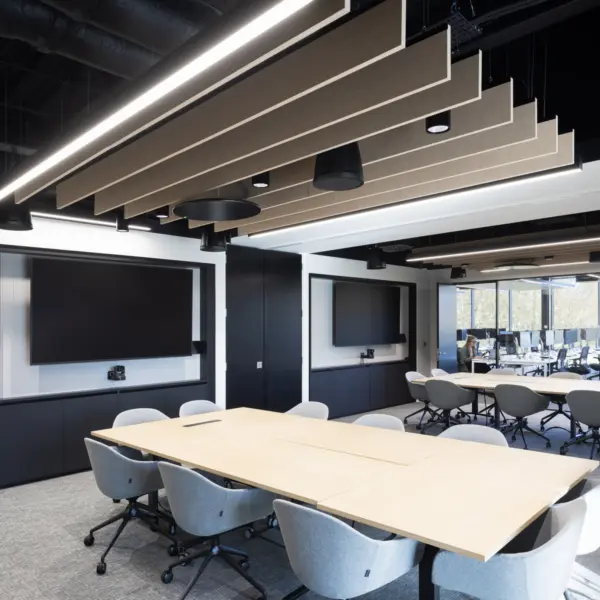A modern meeting room with a large rectangular table surrounded by upholstered swivel chairs. Two large wall‑mounted screens sit on opposite walls above built‑in black cabinetry. Suspended acoustic panels and linear lighting are arranged across the ceiling. A second table with matching chairs is visible in the background near full height windows that look out onto trees.