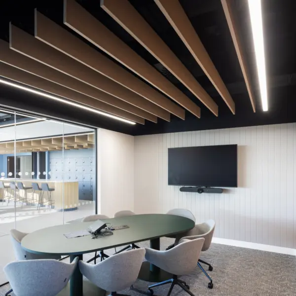 A small meeting room with an oval table surrounded by six upholstered swivel chairs. A large wall‑mounted screen and soundbar sit on a ribbed white feature wall, with an orange accent wall to the right. Suspended acoustic baffles run across the ceiling with linear lights fitted between them. A full height glass wall on the left looks out into an open workspace with a counter and stools visible in the background.