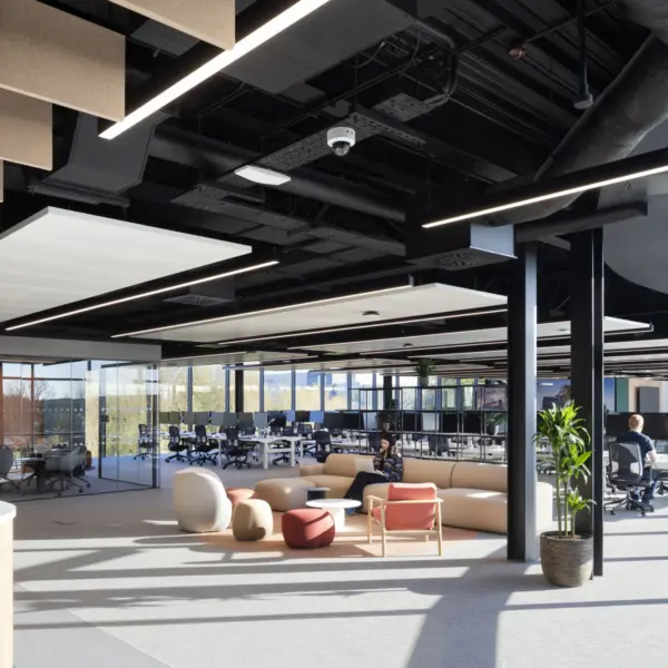 A bright open office area with soft seating in neutral and warm tones arranged near full height windows. Beyond the seating, rows of desks and task chairs fill the main workspace, separated by glass partitions. The ceiling features exposed black services with linear lighting and suspended acoustic panels. A tall plant sits beside a structural column, and a curved counter is partially visible on the left.