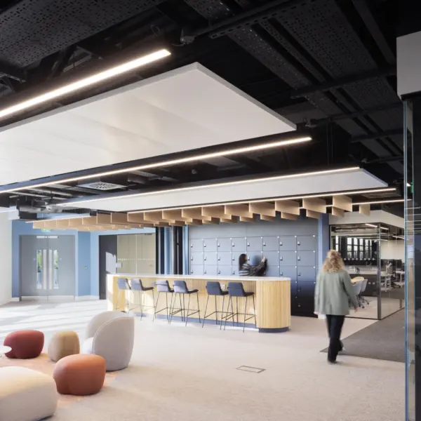 A bright open office space with soft seating in various colours arranged near full height windows. A bar height counter with stools sits in the centre of the room with a wall of lockers behind it. One person stands at the counter and another walks across the space. The ceiling features exposed black panels and linear lighting. A glass meeting room is visible on the right.