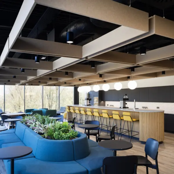 A modern workplace café with a long wooden counter and a row of yellow bar stools facing a dark kitchen backdrop. Round pendant lights hang above the counter. In the foreground, modular blue seating surrounds a central planter filled with greenery, alongside small black tables and chairs. Full height windows line the left side, looking out onto trees, while the ceiling features exposed services and suspended acoustic panels.