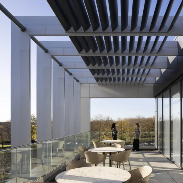 A terrace with round café tables and light chairs arranged along a glass balustrade. The space is partially shaded by a series of horizontal louvres overhead, which cast linear shadows across the floor. Floor to ceiling glazing runs along the right side of the terrace, reflecting the outdoor space. Beyond the terrace, trees and open landscape are visible. Two people stand near the far end of the terrace.