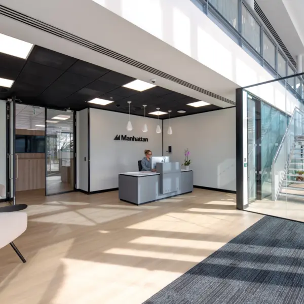 Modern office reception area with light wood flooring, a grey welcome desk and a feature wall displaying the Manhattan logo. A person is seated at the reception desk, while another person walks up a glass-sided staircase in a bright atrium. The space features full-height glazing, white walls, black ceiling panels with integrated lighting and contemporary lounge seating, with natural daylight creating strong shadows across the floor.
