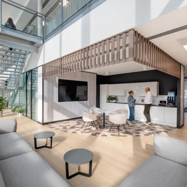 Bright, modern office breakout area with light wood flooring, grey sofas and small round tables. A kitchenette with white cabinetry sits beneath a timber slatted feature structure, while two people stand beside a coffee machine. A large wall-mounted screen, indoor plants and a glass-and-metal staircase leading to an upper level contribute to the open, airy design. Natural light streams through full-height windows along the left side of the space.
