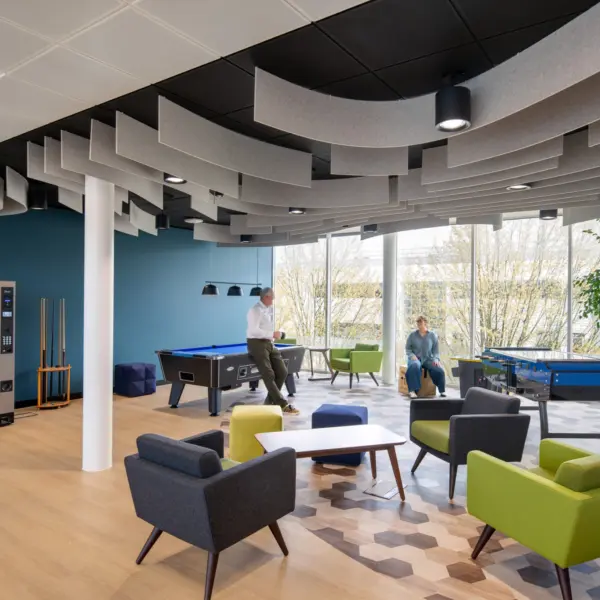 Office breakout and games area with a mix of lounge seating, pool tables and casual meeting spaces. Grey and green armchairs surround low tables on a patterned carpet, while two people are positioned near the games area. Floor-to-ceiling windows along the far wall provide natural daylight and views of trees outside. The space includes a vending machine, cue rack and feature acoustic ceiling baffles in a curved arrangement above, creating a bright and contemporary environment for relaxation and informal collaboration.