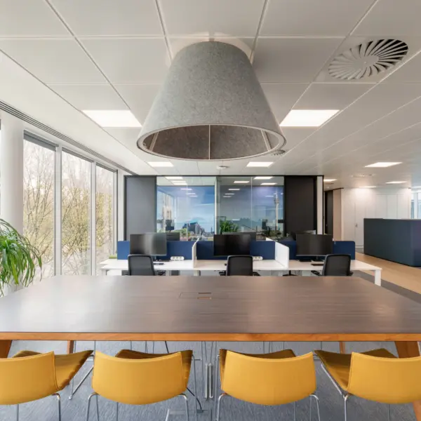 Contemporary open-plan office workspace with a large dark wood collaboration table surrounded by mustard-coloured chairs. A grey acoustic pendant light hangs above the table, while floor-to-ceiling windows along the left side provide natural daylight and views of trees outside. Beyond the meeting area, desks with computer monitors sit within the main workspace, featuring blue privacy screens, light wood flooring and modern finishes throughout. Indoor plants, white structural columns and glazed meeting rooms contribute to the bright, professional environment.