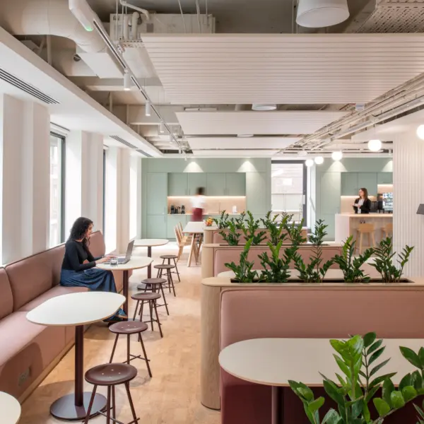 Contemporary office café and breakout area with banquette seating, round tables and integrated planters filled with greenery. Large windows bring natural light into the space, while a kitchenette and refreshment counter sit at the far end. A wall-mounted display screen, soft colour palette and exposed ceiling services create a modern, welcoming environment designed for informal meetings, dining and collaborative work.