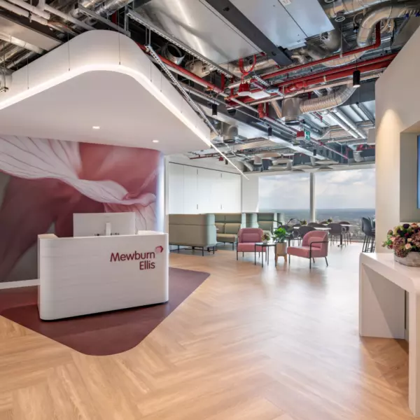A contemporary open-plan office interior featuring a reception area with a sleek white desk bearing the name "Mewburn Ellis" in red lettering and a matching red logo. Behind the desk is a striking wall mural with an abstract pink and white design. The ceiling showcases exposed ductwork and piping, contributing to an industrial aesthetic. To the right, a seating area includes pink and grey upholstered chairs arranged around small circular tables, set beside large windows that provide ample natural light and a broad external view. A flat-screen television is mounted on the adjacent wall above a console table decorated with a vase of flowers. The floor is laid with light wood in a herringbone pattern, adding a touch of warmth and texture to the space.