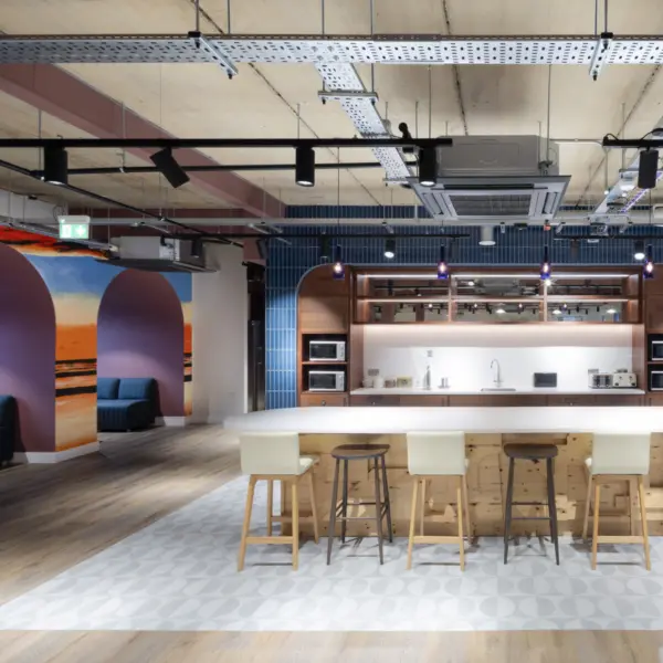 Wide view of an open plan breakout space with timber flooring, exposed ceiling services and suspended lighting. Soft seating and small tables sit in the foreground, leading towards a communal kitchen with a white island and stools at the back. A pool table and wall mural with arched forms and a sunset scene appear on the left, while booth seating and armchairs define informal meeting areas on the right. Provide your feedback on BizChat