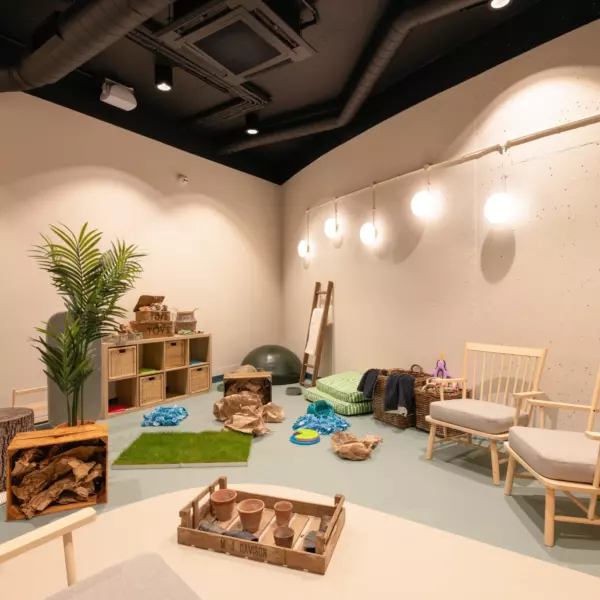 Bright enrichment room with a playful, natural theme. Light walls and soft flooring in green and beige tones set a calming backdrop. The space includes wooden shelving filled with textured play items, faux grass mats, and natural elements like logs and stones. Pendant globe lights and simple wooden chairs complete the relaxed, creative environment.