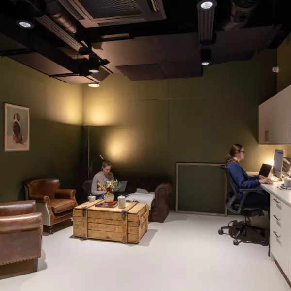 Cosy staff office with deep green walls and contrasting white cabinetry. The space combines functionality with comfort, featuring leather armchairs, a rustic wooden coffee table, and framed dog-themed artwork. Overhead black ceiling panels and spotlights create a modern, slightly industrial aesthetic.