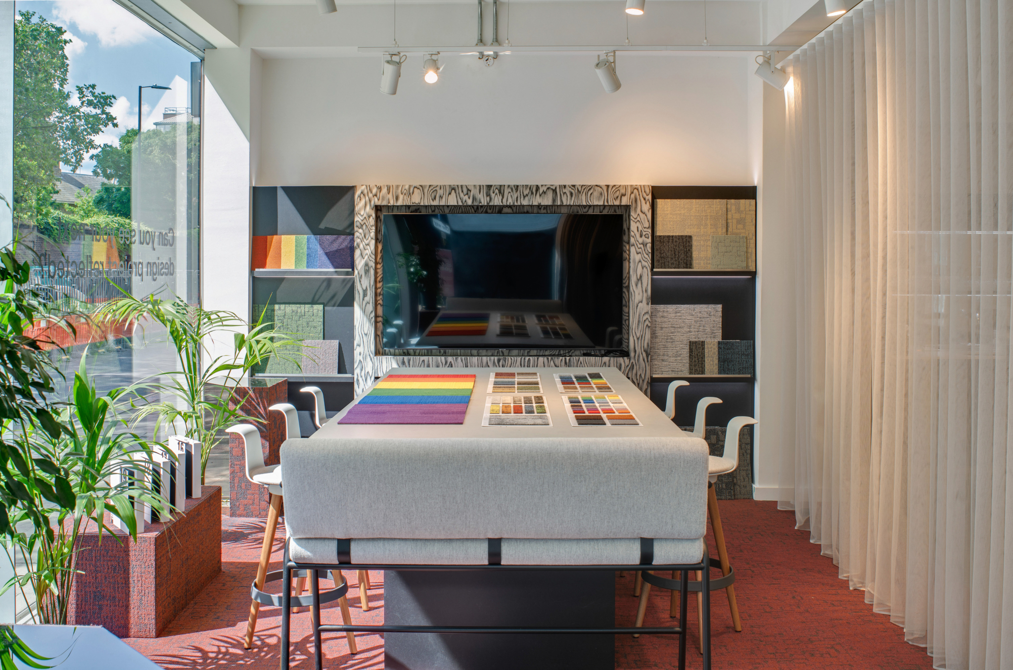 An interior view of a design studio or showroom featuring a large central table displaying an array of colour samples and material swatches. Chairs are arranged around the table, and potted plants line the left side of the space. A large mirror or screen on the back wall reflects additional samples. The room is illuminated by natural light streaming through expansive windows on the left, enhancing the clean and curated layout.
