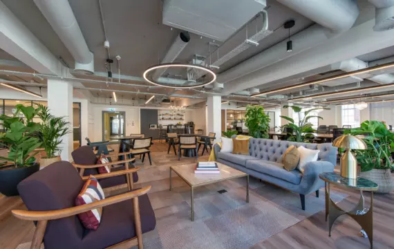 A modern open plan office space featuring two purple armchairs with wooden frames and colorful cushions, a gray tufted sofa with pillows and a coffee table with books. A small round side table with a gold lamp sits beside the sofa. The background includes additional seating areas with tables and chairs, green plants and large windows letting in natural light. The ceiling has an industrial design with exposed ducts and pipes.