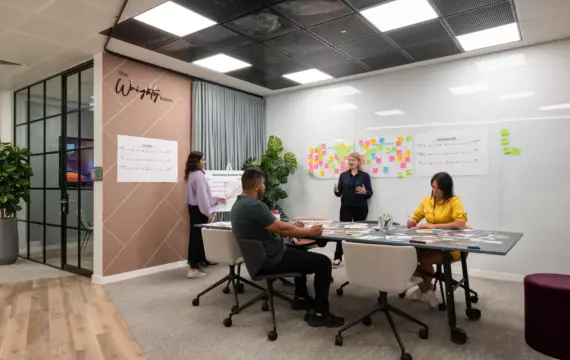 Four people in a modern office meeting room collaborating on a project. One person stands near a wall covered with sticky notes and charts presenting to the group. The others are seated around a large table with papers and materials spread out. The room features glass walls, mixed flooring, potted plants and ceiling lights.