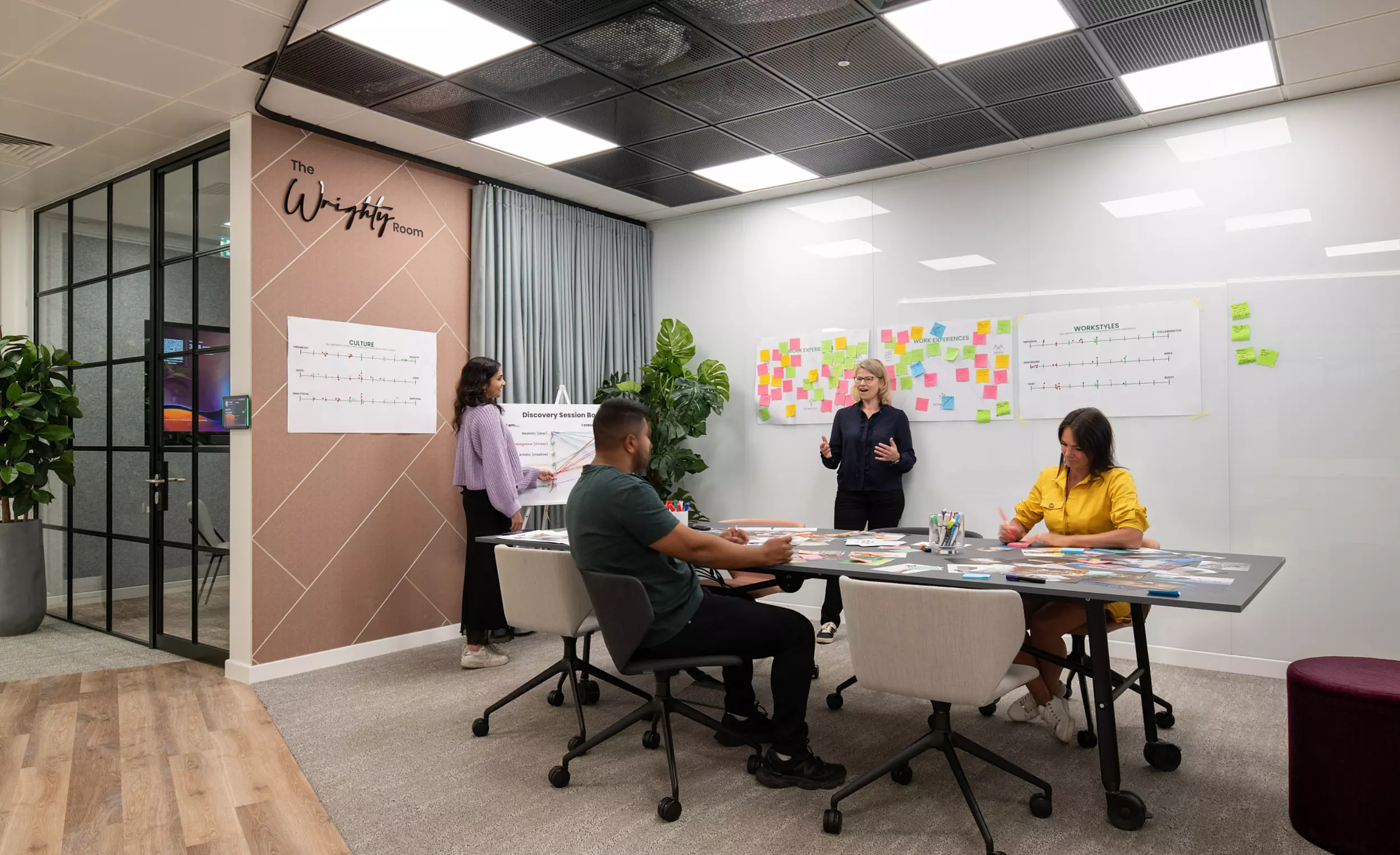Four people in a modern office meeting room collaborating on a project. One person stands near a wall covered with sticky notes and charts presenting to the group. The others are seated around a large table with papers and materials spread out. The room features glass walls, mixed flooring, potted plants and ceiling lights.