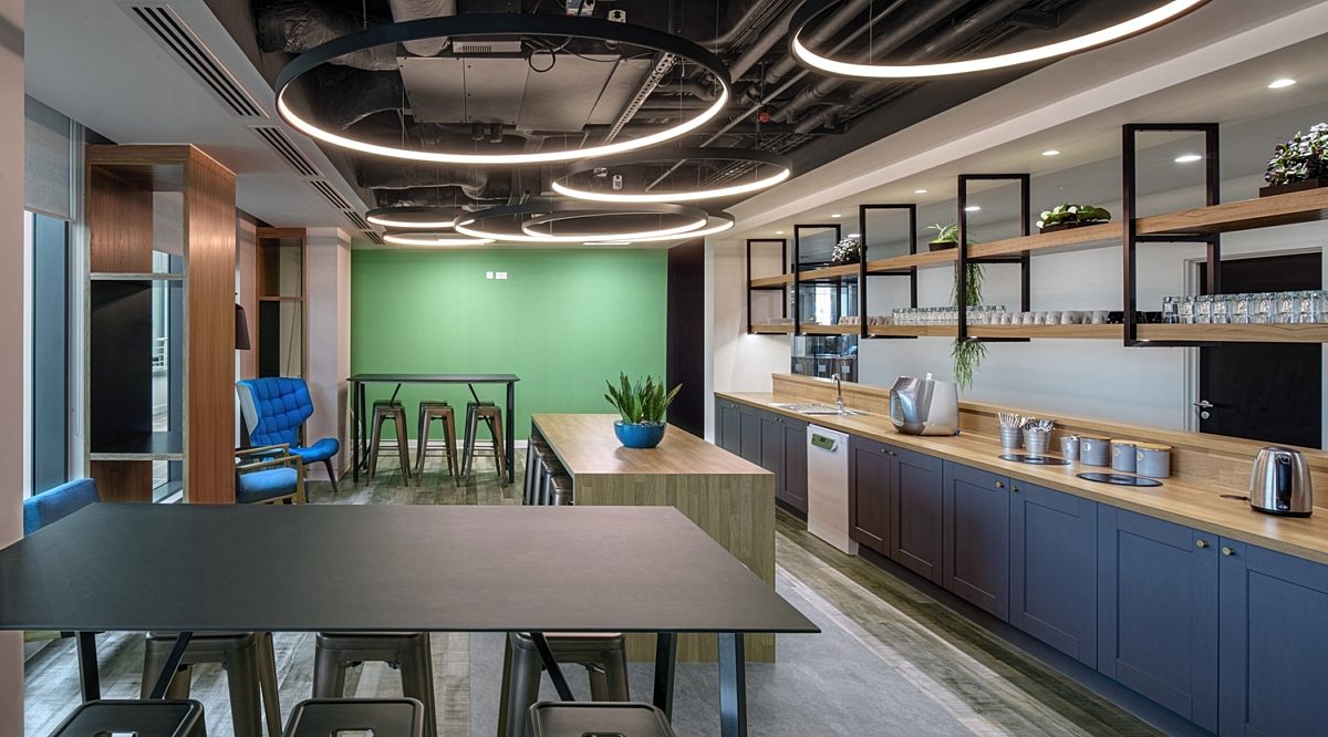 A chic office kitchen and breakout area with circular overhead lights, a green accent wall, contemporary furniture, and wooden shelving