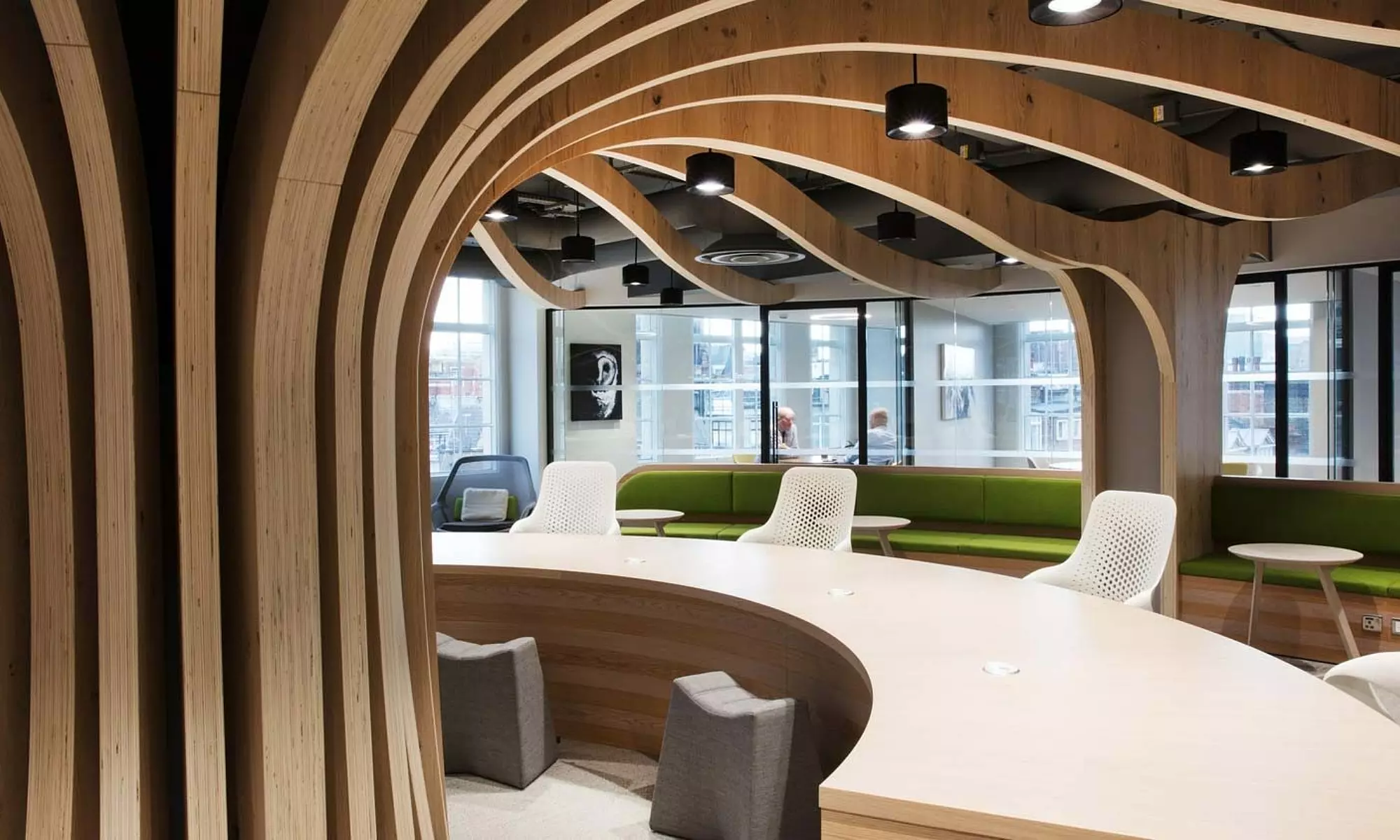 An office space with distinctive architectural features, including curved wooden ceiling beams and large windows that allow natural light to enter. The room is furnished with green cushioned bench seating along the walls, white chairs arranged around a curved table, and small grey stools. Black pendant lights hang from the ceiling, contributing to the room’s cohesive and thoughtfully designed aesthetic.