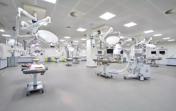 A spacious operating theatre featuring multiple surgical tables surrounded by advanced medical equipment. The ceiling is fitted with bright, ceiling-mounted lighting and several articulated surgical lights. Monitors and medical devices are strategically placed around the room for accessibility. The floor is spotless, and the walls are lined with built-in cabinets and storage units, contributing to a highly functional and organised clinical environment.