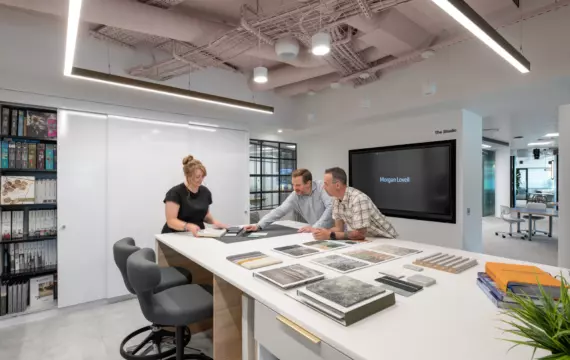 A well-lit room with three individuals gathered around a white table, examining an assortment of fabric swatches and samples. The space features open shelving along the walls, filled with books and decorative objects. The overall layout is clean and thoughtfully arranged, supporting collaborative work and creative exploration.