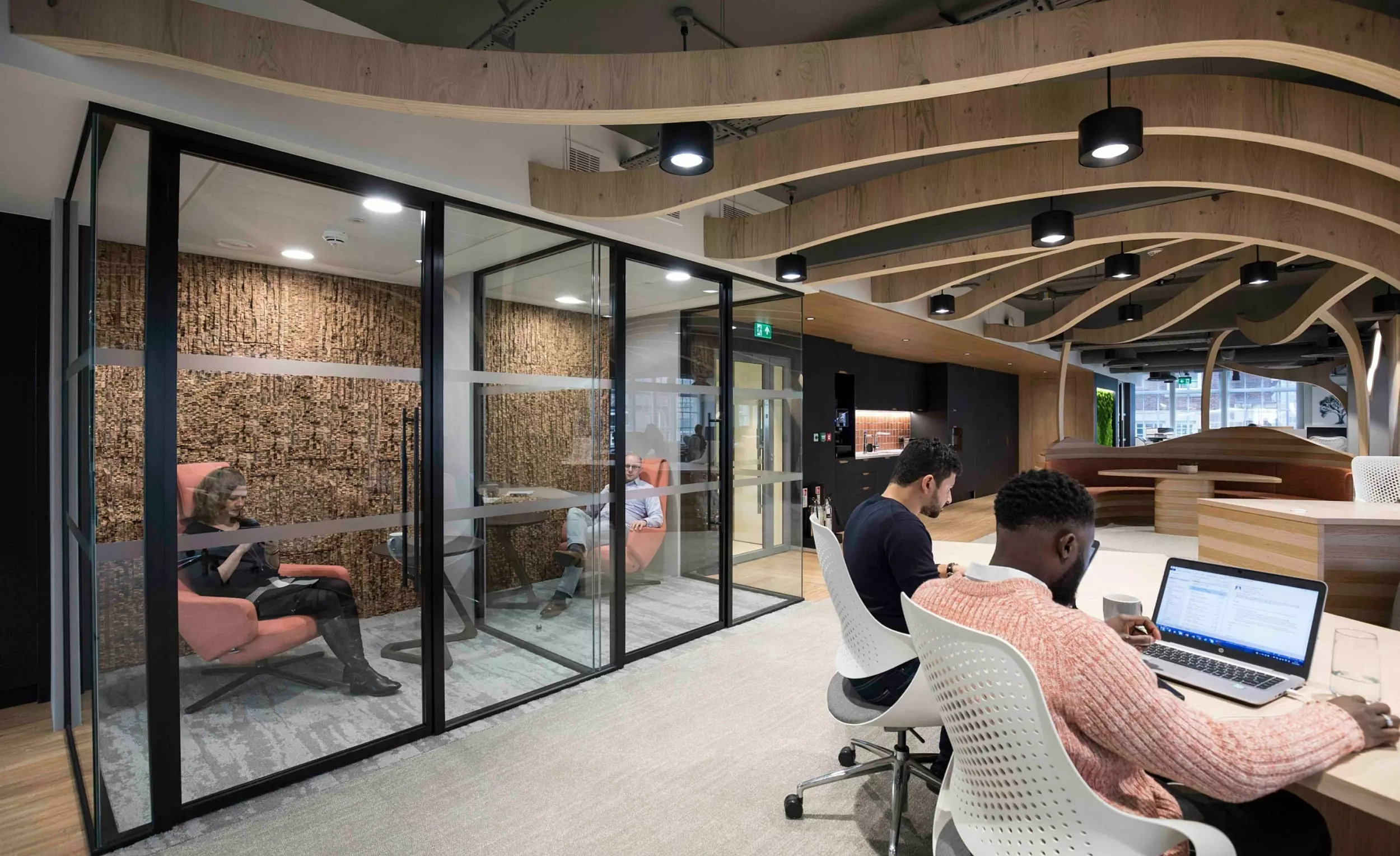 An open-plan office space featuring two individuals working on laptops at a shared desk. The background includes a glass-enclosed meeting room with one person seated inside. The space is characterised by wooden ceiling beams, clean lines, and contemporary furnishings, creating a professional and collaborative environment.