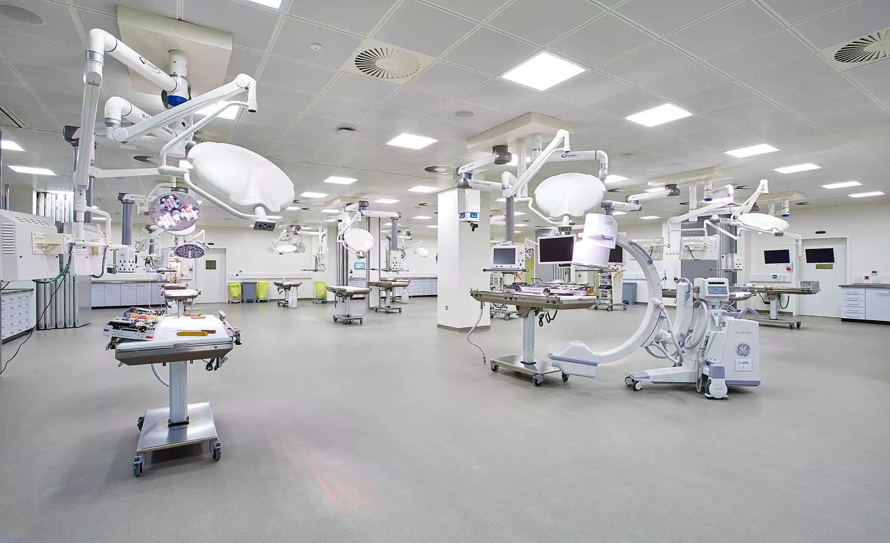 A spacious operating theatre featuring multiple surgical tables surrounded by advanced medical equipment. The ceiling is fitted with bright, ceiling-mounted lighting and several articulated surgical lights. Monitors and medical devices are strategically placed around the room for accessibility. The floor is spotless, and the walls are lined with built-in cabinets and storage units, contributing to a highly functional and organised clinical environment.