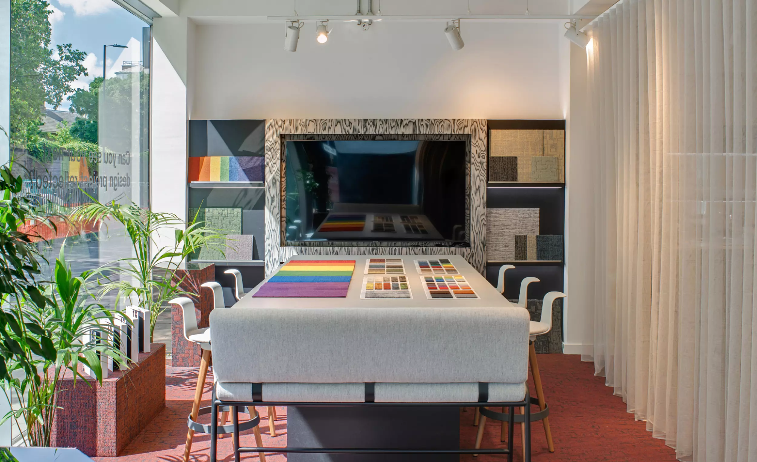 An interior view of a design studio or showroom featuring a large central table displaying an array of colour samples and material swatches. Chairs are arranged around the table, and potted plants line the left side of the space. A large mirror or screen on the back wall reflects additional samples. The room is illuminated by natural light streaming through expansive windows on the left, enhancing the clean and curated layout.
