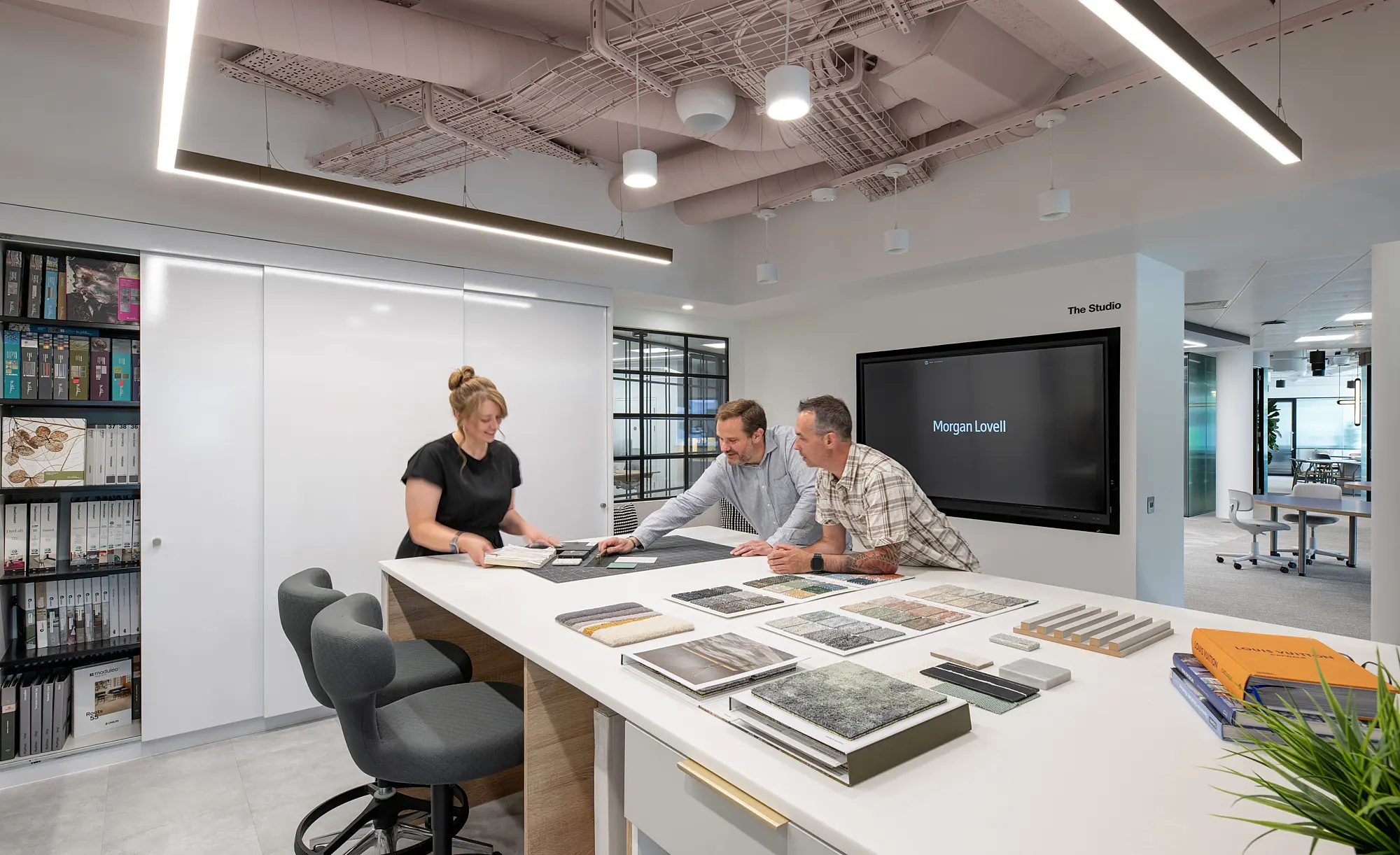 A well-lit room with three individuals gathered around a white table, examining an assortment of fabric swatches and samples. The space features open shelving along the walls, filled with books and decorative objects. The overall layout is clean and thoughtfully arranged, supporting collaborative work and creative exploration.