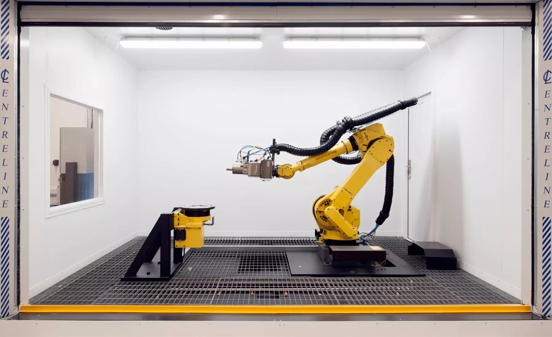 A yellow industrial robotic arm situated within a white, enclosed workspace. The arm is mounted on a black platform and features a tool attachment at its end. The environment is clean and minimal, with smooth surfaces and a focus on precision and containment.