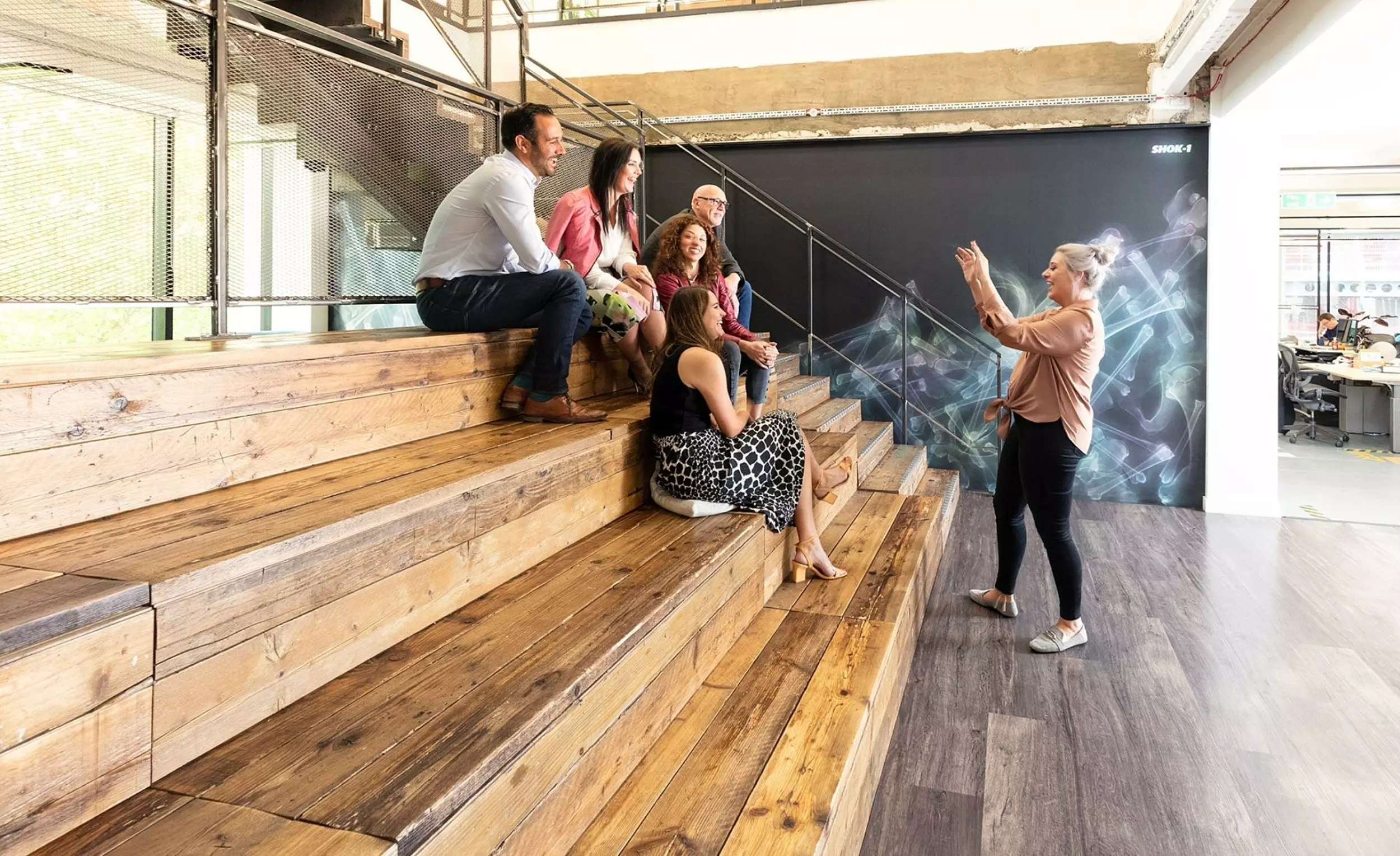 An office setting featuring five individuals, with four seated on wooden bleacher-style steps and one person standing. The background includes a black wall with an abstract design and an open-plan workspace with desks and chairs. The layout supports informal presentations and collaborative discussions within a flexible environment.