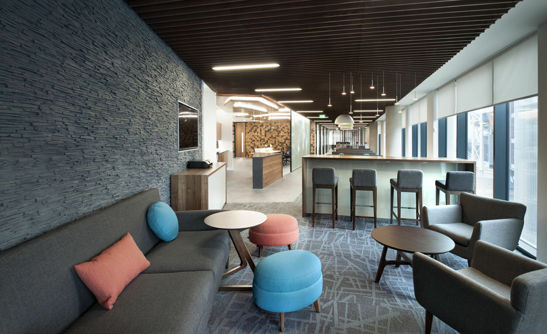 An open office area with textured grey walls, wood accented ceiling, sleek furniture in cool tones, and bright natural light from floor-to-ceiling windows