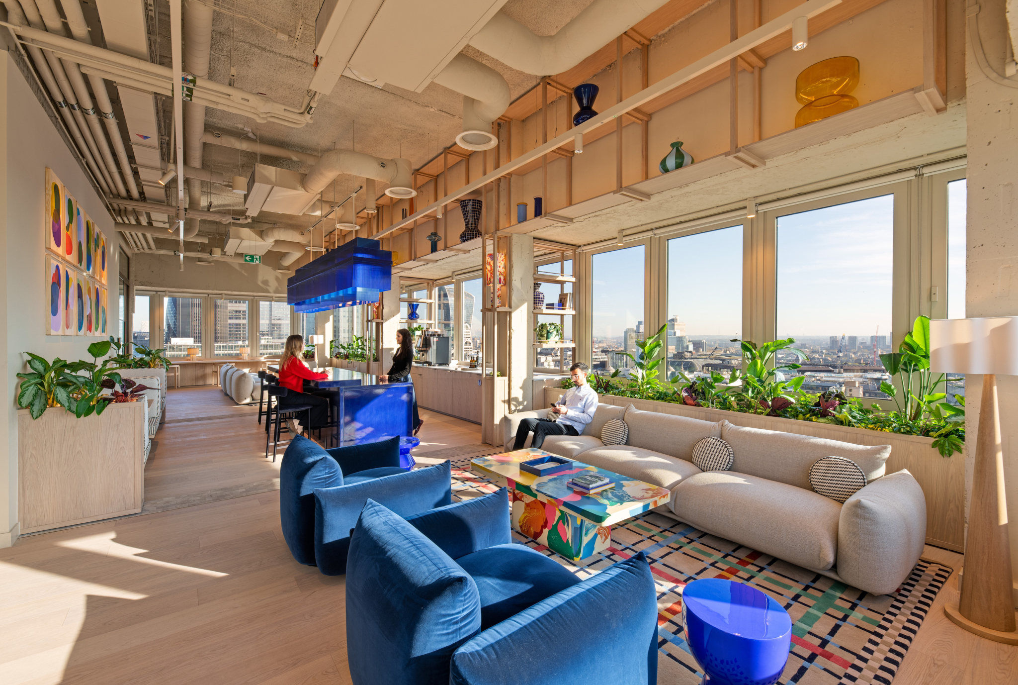 Contemporary office lounge with panoramic views across the London skyline through floor-to-ceiling windows. The space features a seating area with a curved beige sofa, four blue lounge chairs and a colourful coffee table set on a patterned rug. A glossy blue bar with matching pendant lighting sits to the left, while open timber shelving displays decorative glassware and integrated planters filled with greenery. Natural daylight floods the interior, complementing the light wood finishes, exposed ceiling services and vibrant design details throughout the workplace.