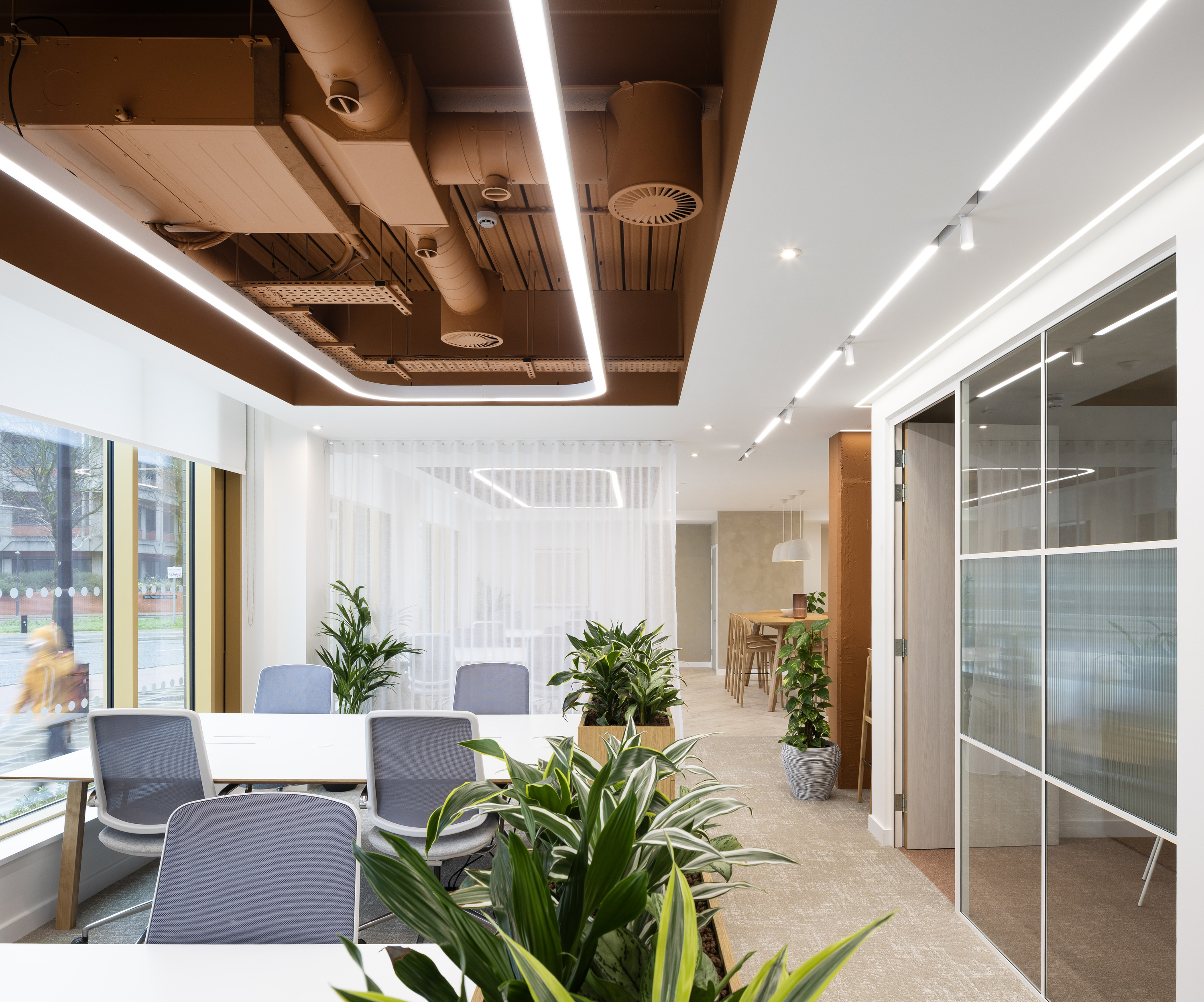 Modern office interior with a clean, minimalist design. In the foreground, a white meeting table is surrounded by several light grey office chairs, with multiple green potted plants placed around the seating area. Large windows on the left let in natural light and show a street scene outside. The ceiling features exposed ductwork painted in a warm brown tone, contrasted with linear LED light strips. In the background, sheer white curtains partially divide the space, leading to a casual breakout area with high wooden stools and a counter. On the right side, a glass-partitioned room with a sliding door is visible. The overall space uses a mix of neutral colours, warm wood accents and indoor greenery to create a bright, contemporary workplace.