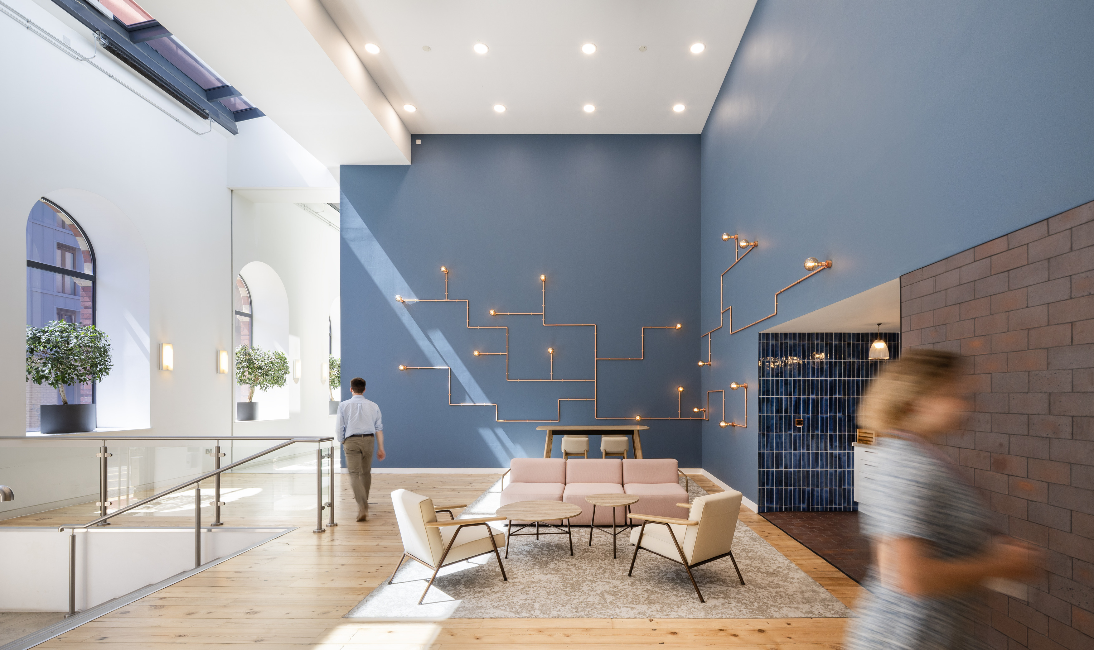 A modern interior space with high ceilings and large arched windows that fill the room with natural light. The seating area features pink and beige furniture on a light rug, set against a blue wall with geometric light fixtures. Potted plants and soft colours create a relaxed, contemporary atmosphere.